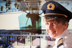 Female airline pilot wearing uniform with epauletes, hat with golden wings sitting inside airliner with visible cockpit during flight.